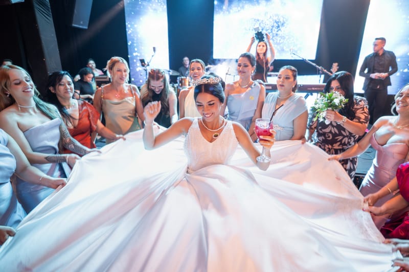 Noiva rindo na pista, cercada pelas amigas que seguram a saia do vestido