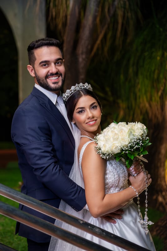 Noivos abraçados e sorrindo à noite, ela com o buquê de rosas brancas