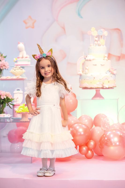 Menina de vestido branco ao lado da mesa de doces com tema de unicórnio